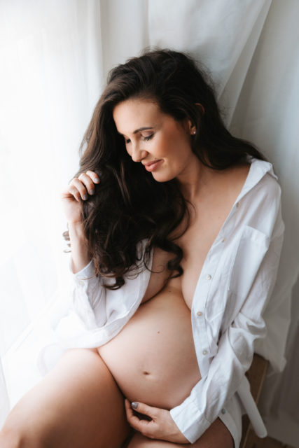 portrét těhotné ženy sedící v okně u bílé záclony